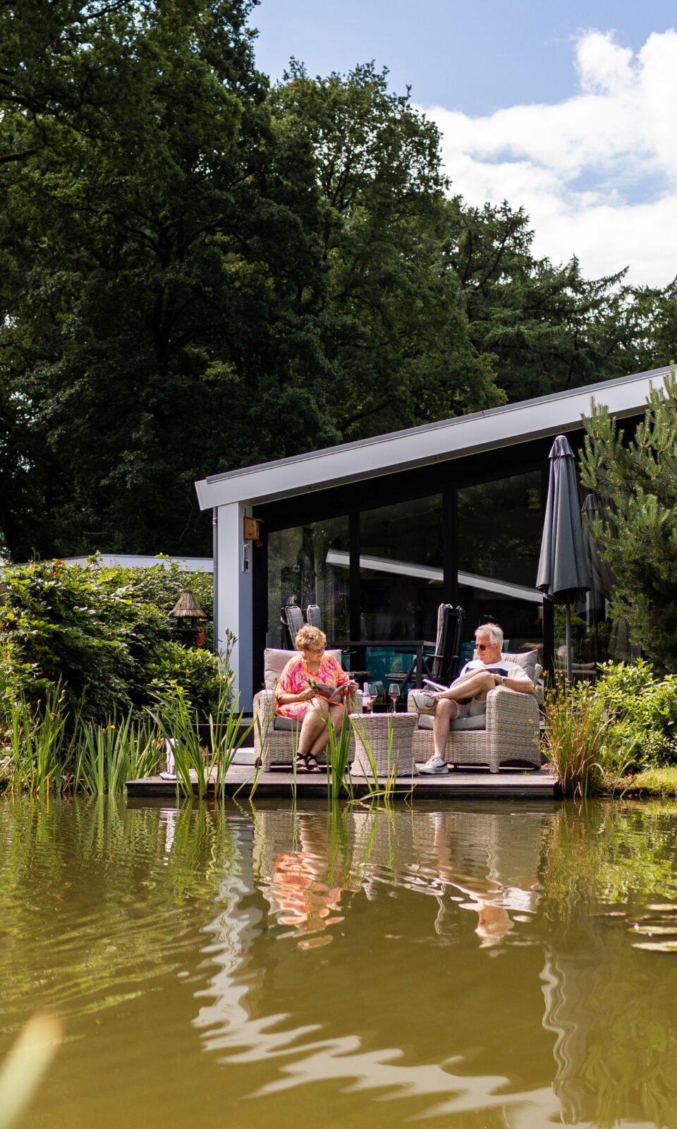 Vakantiehuisje aan het water, op het vlonder zit een echtpaar een boekje te lezen en een wijntje te drinken