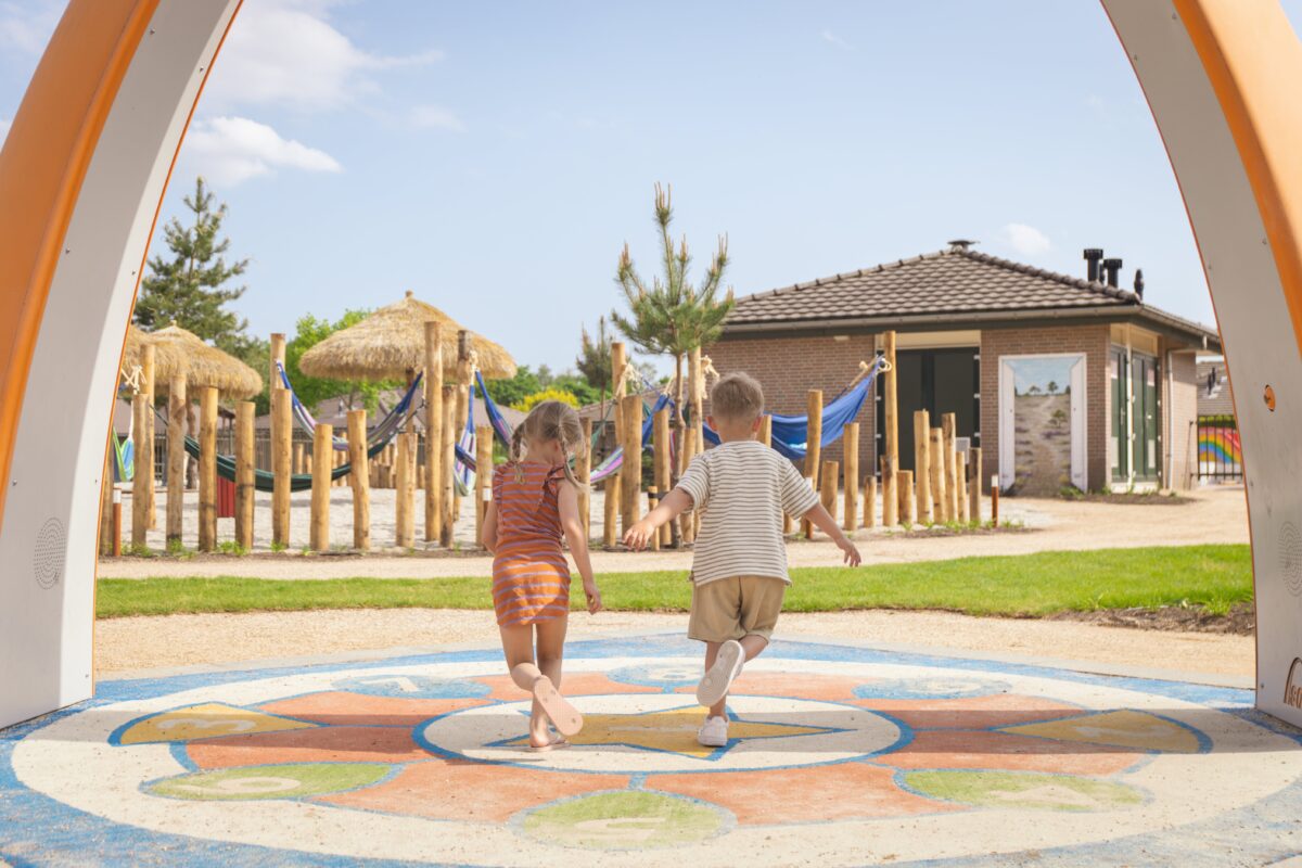 Twee kinderen rennen onder een boog door richting een houten speeltuin en een houten huisje