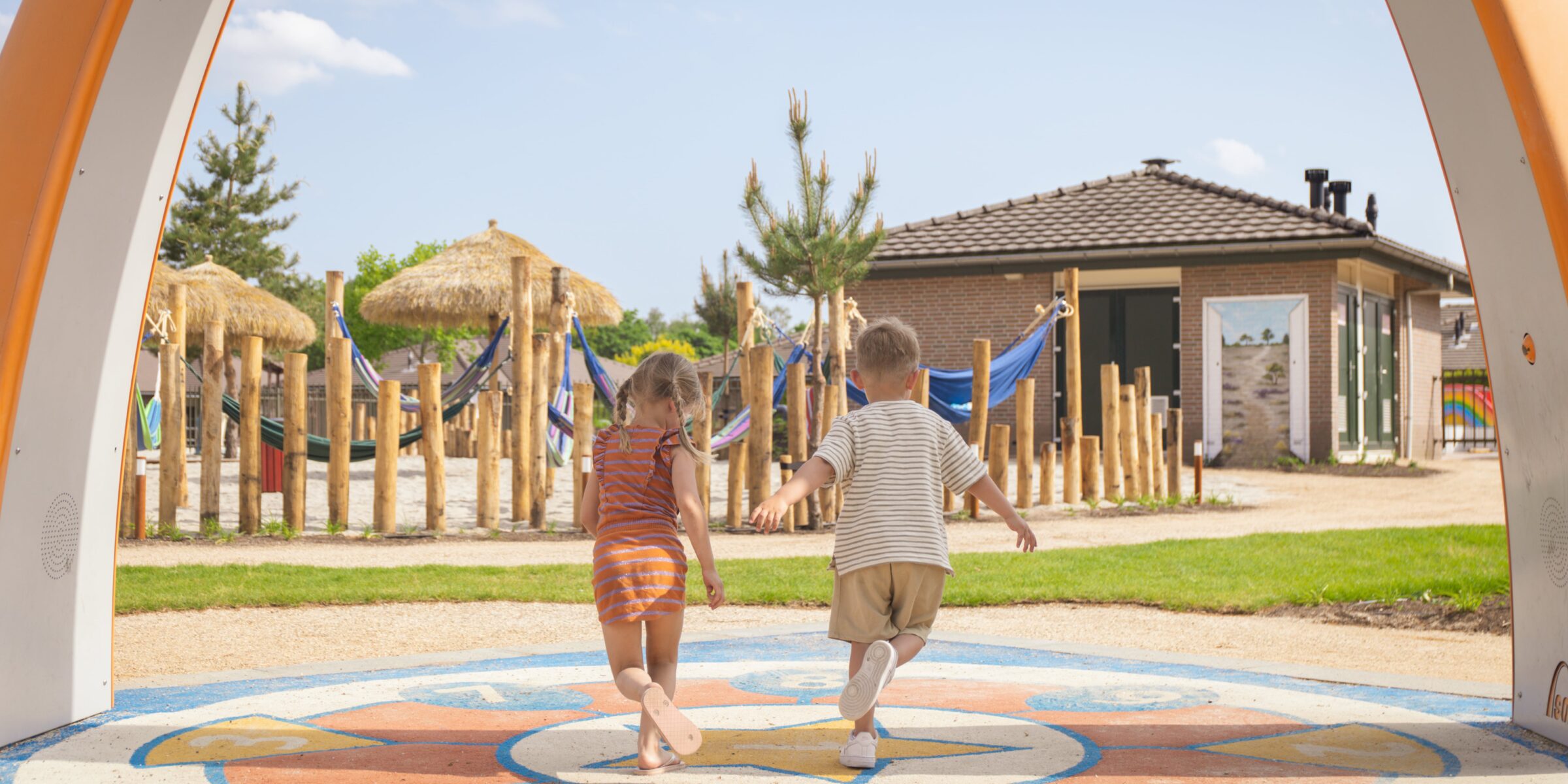 Twee kinderen rennen onder een boog door richting een houten speeltuin en een houten huisje