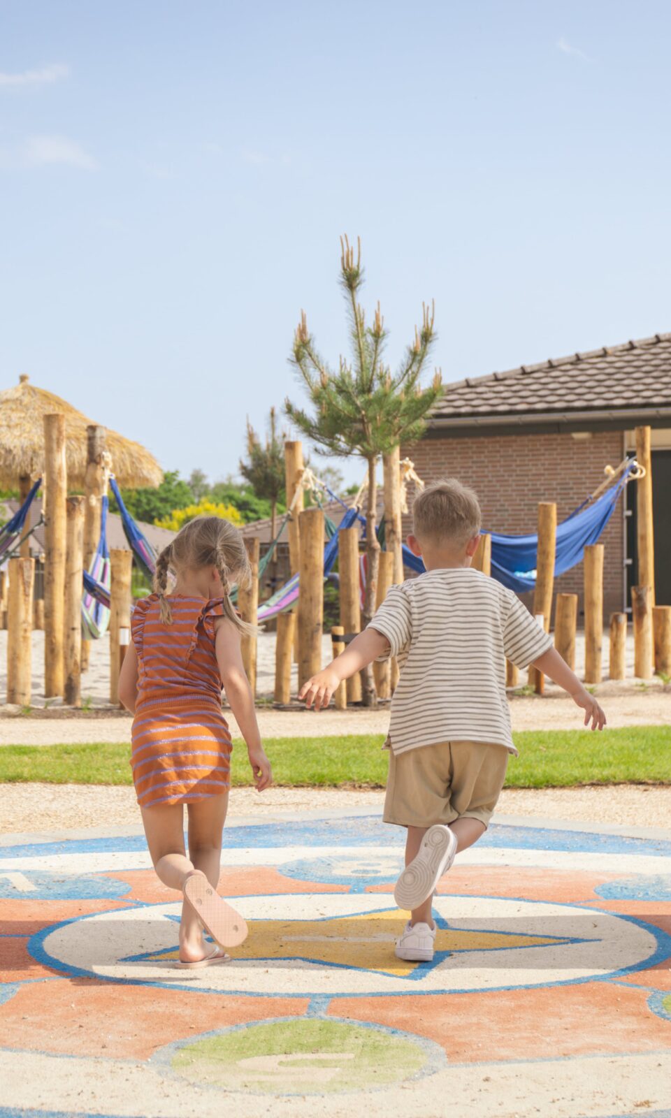 Twee kinderen rennen onder een boog door richting een houten speeltuin en een houten huisje