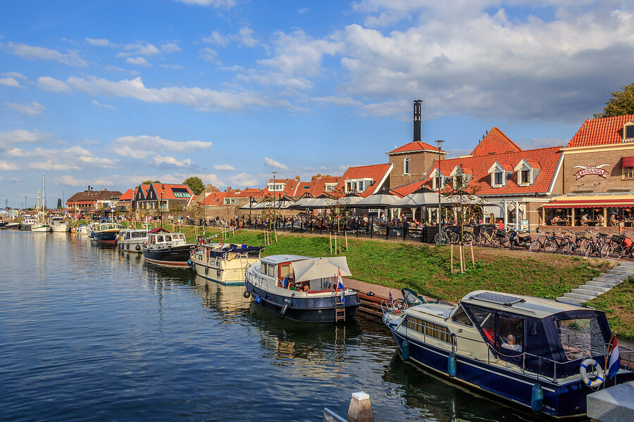 Een rij boten aangemeerd aan de kade. Op de kade is een terras vol met mensen. Op de achtergrond die je huizen en veel fietsen.