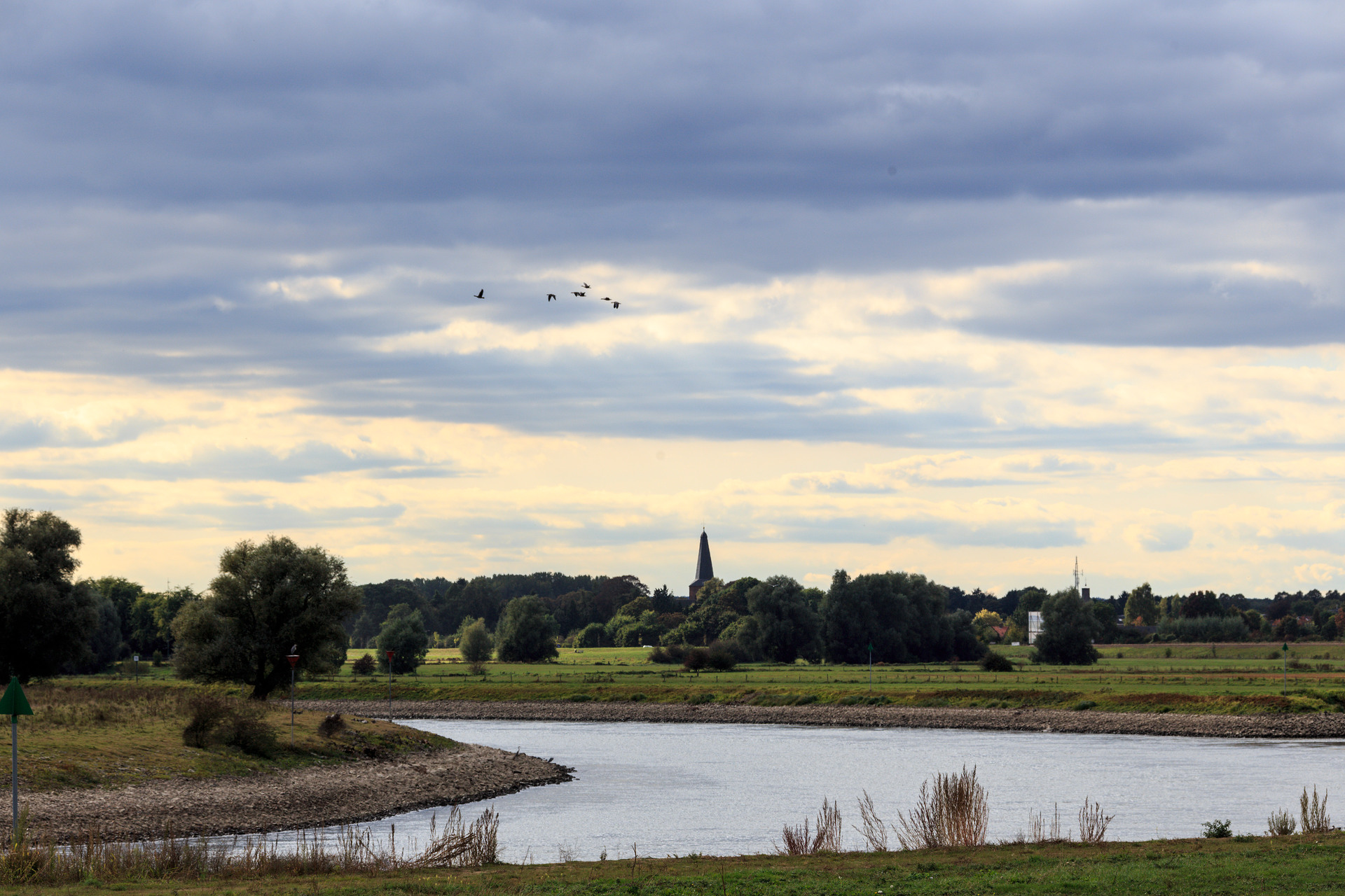 Rivier door het landschap. Op de achtergrond een kerk. In de lucht vliegen vogels en zie je de wolken