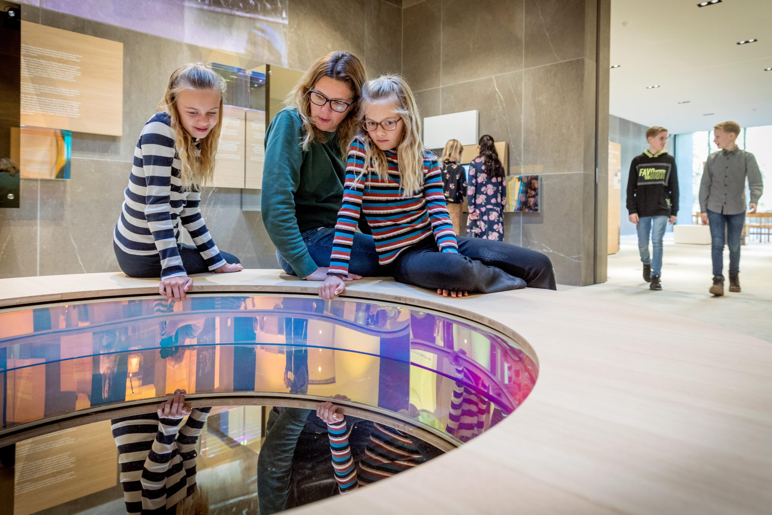 Drie kinderen staan gebogen over een paneel bij het bezoekerscentrum van Ereveld Loenen