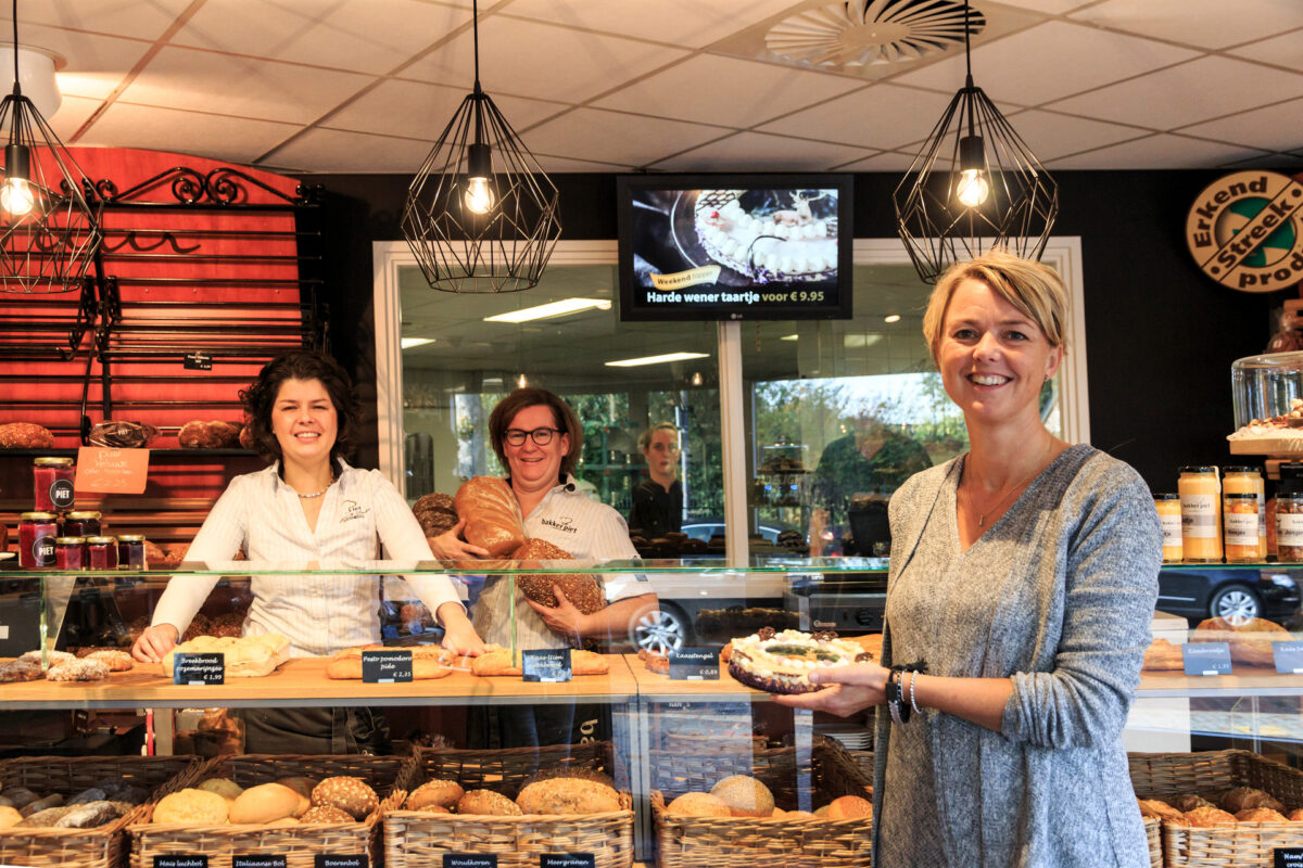 Twee vrouwen poseren voor bakker Piet in Elspeet, met een assortiment brood zichtbaar in de winkel.