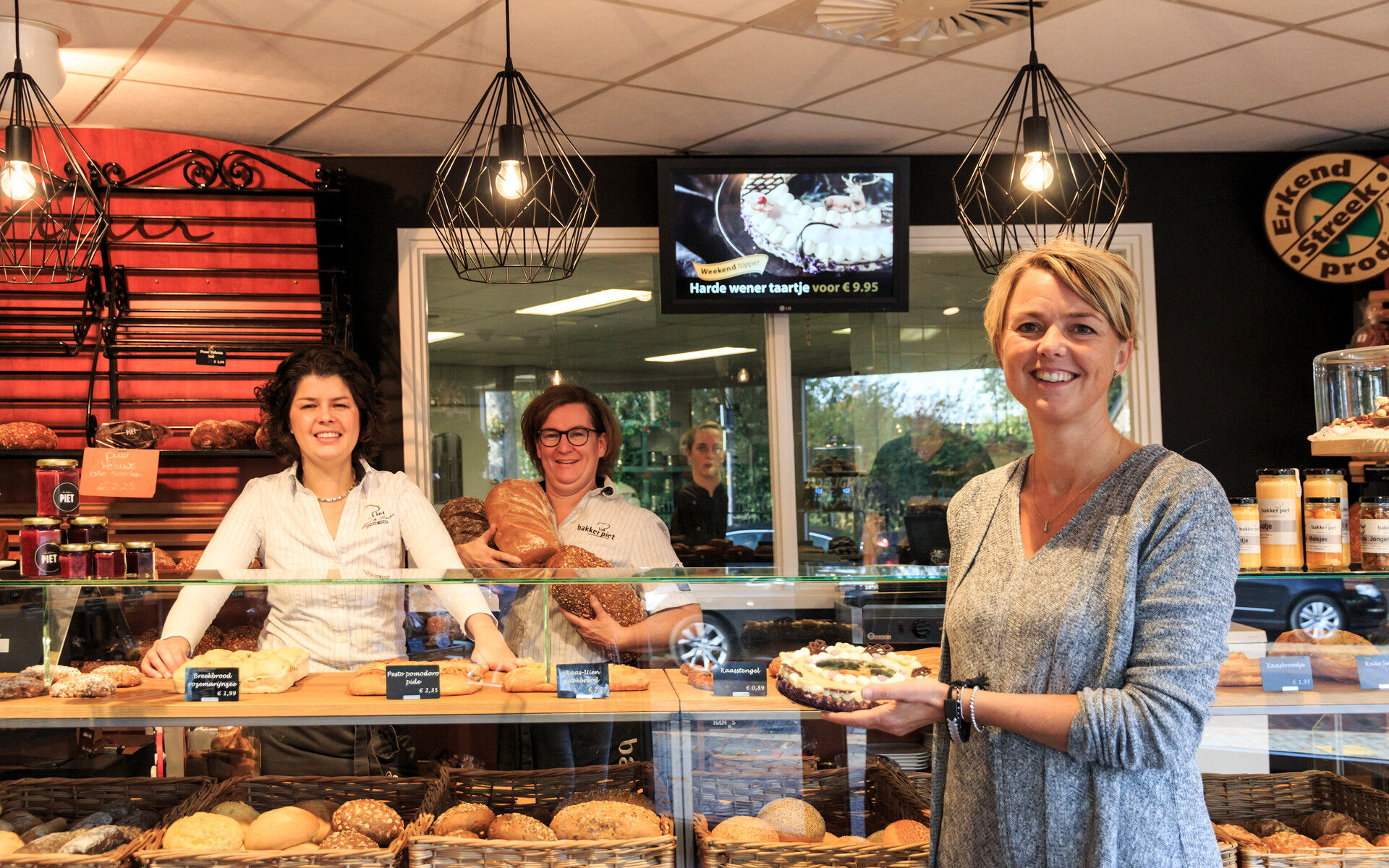 Twee vrouwen poseren voor bakker Piet in Elspeet, met een assortiment brood zichtbaar in de winkel.