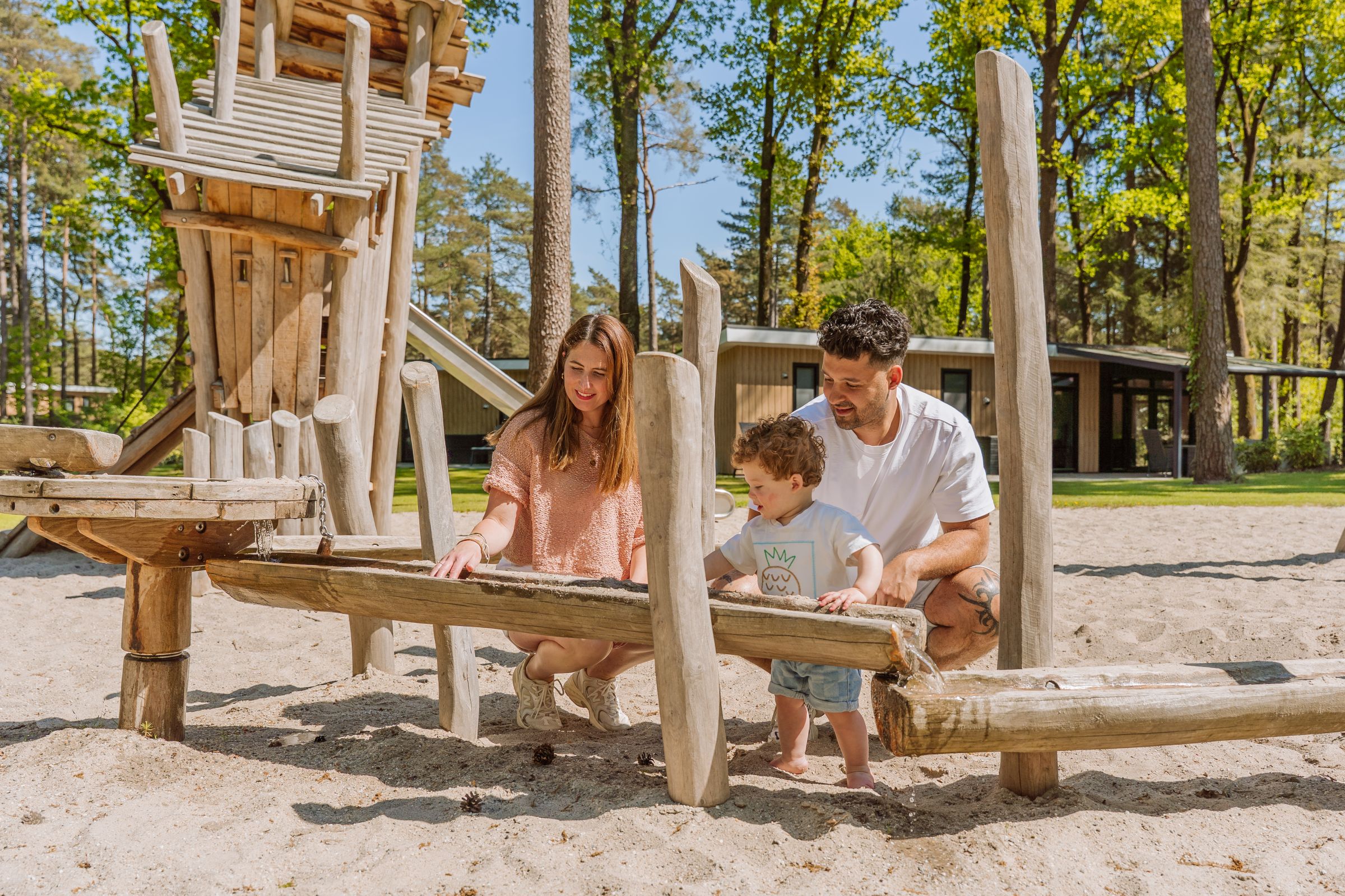 Ouders met klein kind spelen bij een speeltoestel in het zand met een chalet op de achtergrond