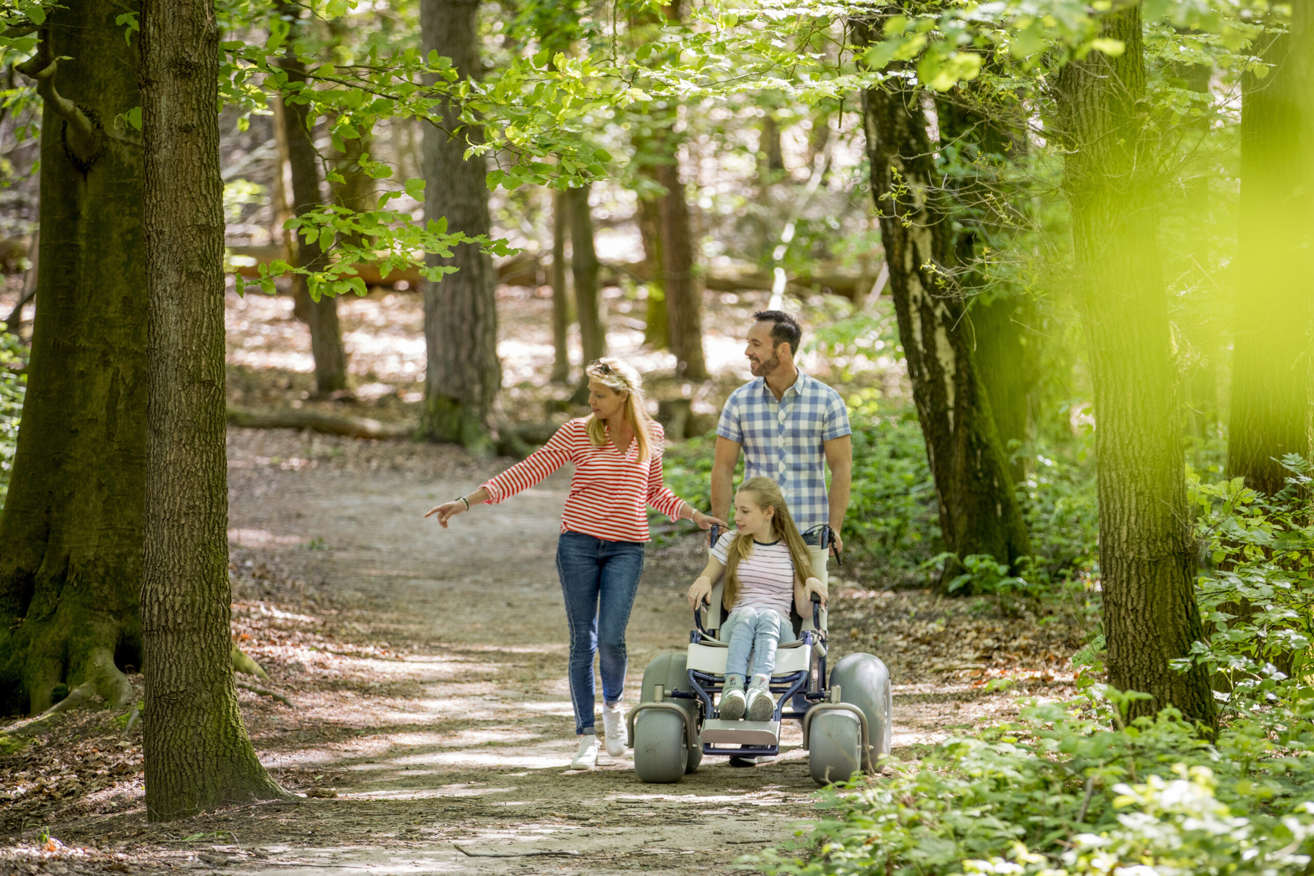 man en vrouw lopen met meisje in rolstoel met dikke banden in een bosrijke omgeving